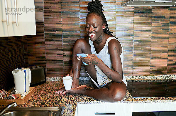 Lächelnde Hipster-Frau  die ein Smartphone benutzt  während sie zu Hause auf dem Küchentisch sitzt