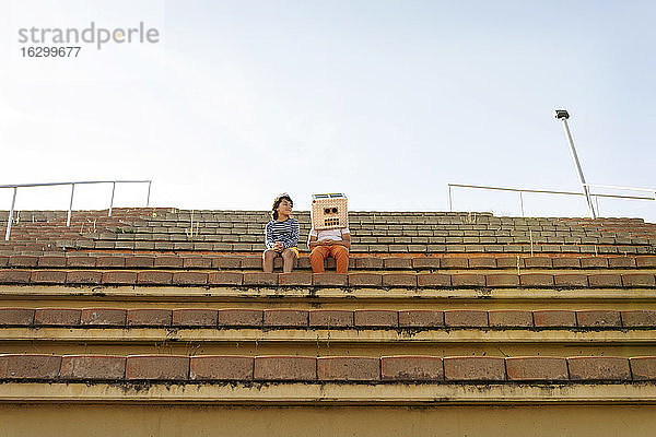 Junge mit Robotermaske sitzt mit einem Freund auf einer Treppe gegen den Himmel an einem sonnigen Tag