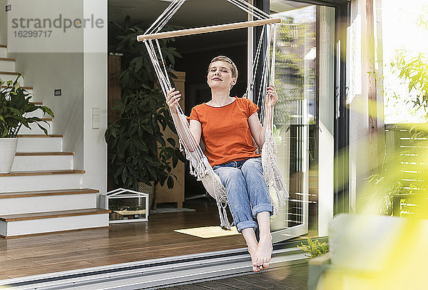 Mittlere erwachsene Frau mit geschlossenen Augen  die sich auf einer Schaukel vor einem Haus auf einer Veranda entspannt