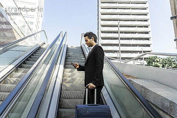 Geschäftsmann mit Koffer  der ein Smartphone benutzt  während er auf einer Rolltreppe in der Stadt steht