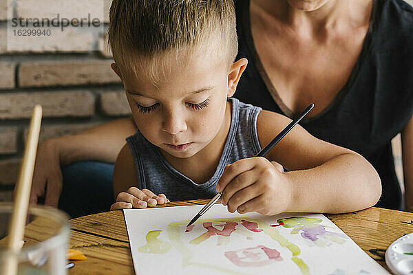Junge malt auf Papier mit Mutter im Hintergrund zu Hause