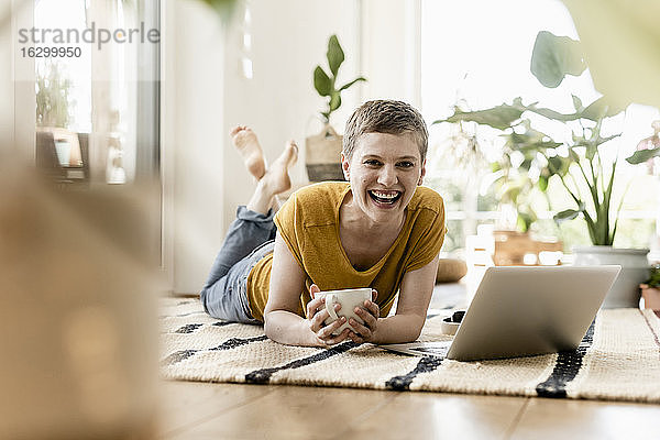 Fröhliche Frau hält eine Kaffeetasse  während sie einen Laptop auf einem Teppich zu Hause benutzt
