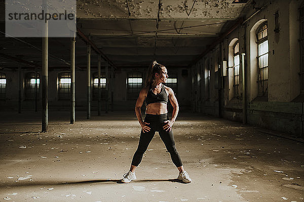 Muskulös gebaute Frau steht mit der Hand auf der Hüfte in einer verlassenen Fabrik
