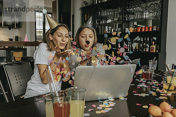 Fröhliche Freunde  die bei einer Videokonferenz über einen Laptop Konfetti ausblasen  während sie zu Hause sitzen