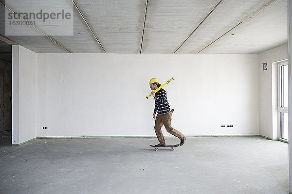 Junger Bauarbeiter  der in einem leeren Haus Skateboard fährt