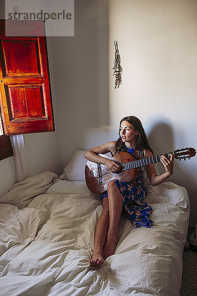 Junge Frau schaut weg  während sie zu Hause auf dem Bett Gitarre übt