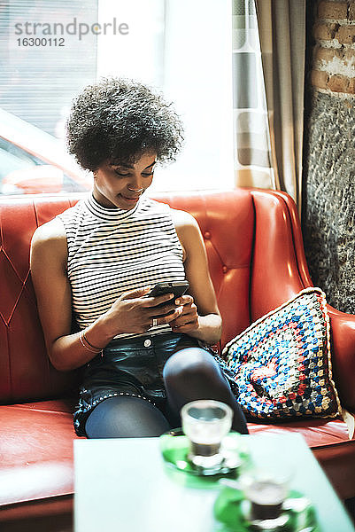Frau mit lockigem Haar  die ihr Smartphone benutzt  während sie sich auf dem Sofa vor dem Fenster in einem Café entspannt