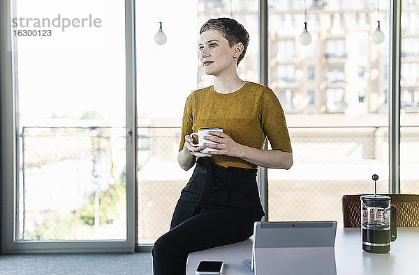 Geschäftsfrau sitzt auf einem Schreibtisch im Büro und hält einen Kaffeebecher