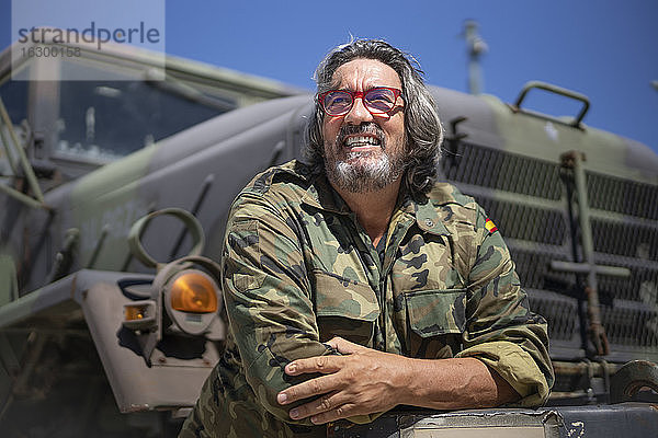 Lächelnder Armeesoldat mit gekreuzten Armen  der sich an einen Militärlastwagen lehnt  an einem sonnigen Tag