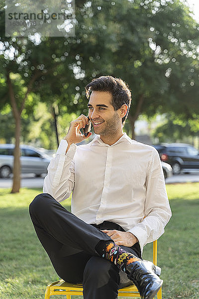 Lächelnder Geschäftsmann  der über sein Smartphone spricht  während er auf einem Stuhl vor einem Baum sitzt