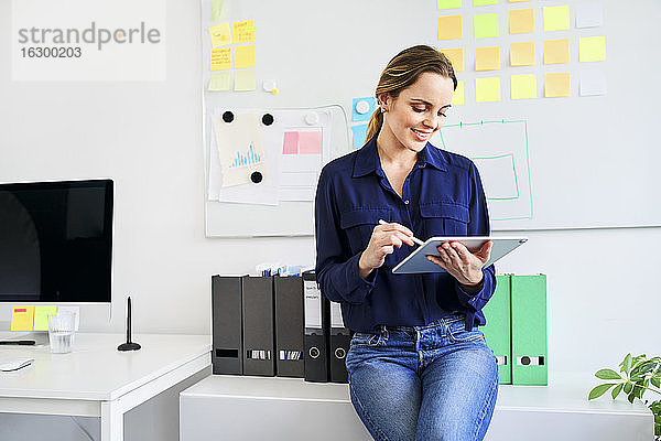 Lächelnde kreative Geschäftsfrau  die ein digitales Tablet benutzt  während sie sich auf einen Schreibtisch im Büro stützt