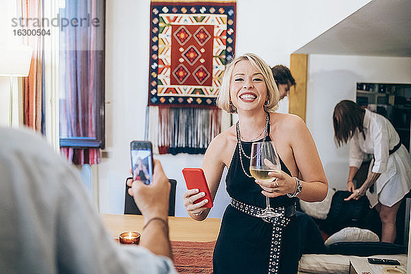 Mann fotografiert fröhliche Freundin  die ihr Smartphone hält  während sie auf einer Party Wein trinkt