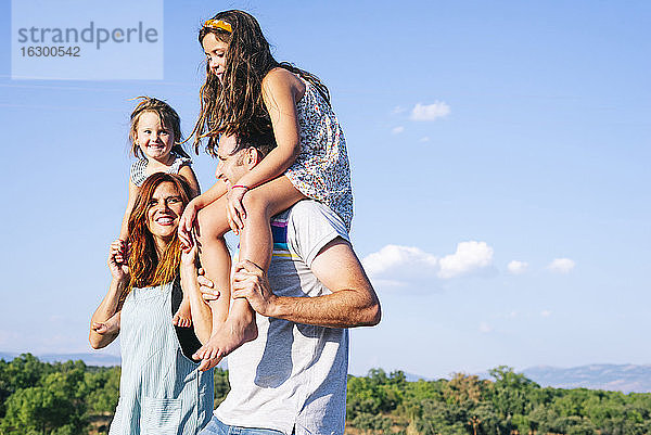 Glückliche Eltern tragen ihre Töchter auf den Schultern gegen den Himmel an einem sonnigen Tag