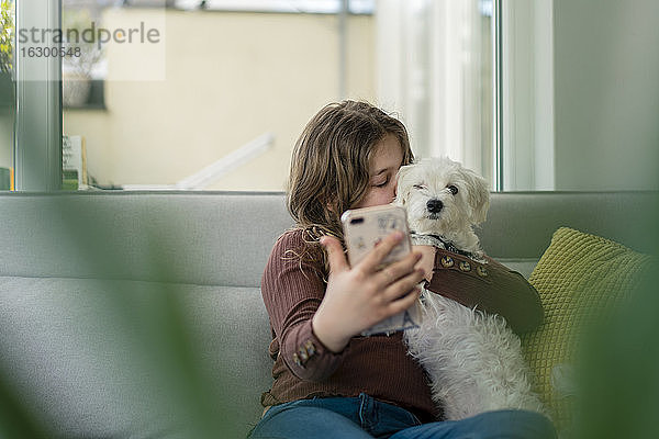 Mädchen  das ein Selfie mit dem Smartphone macht  während es den Hund auf dem Sofa küsst