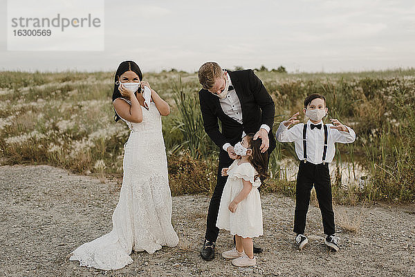 Eltern mit Kindern im Hochzeitskleid tragen einen Mundschutz  während sie während COVID-19 auf einem Feld stehen