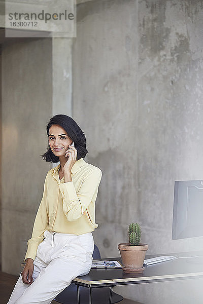 Lächelnde Geschäftsfrau  die über ein Mobiltelefon spricht  während sie auf einem Schreibtisch an der Wand im Büro sitzt