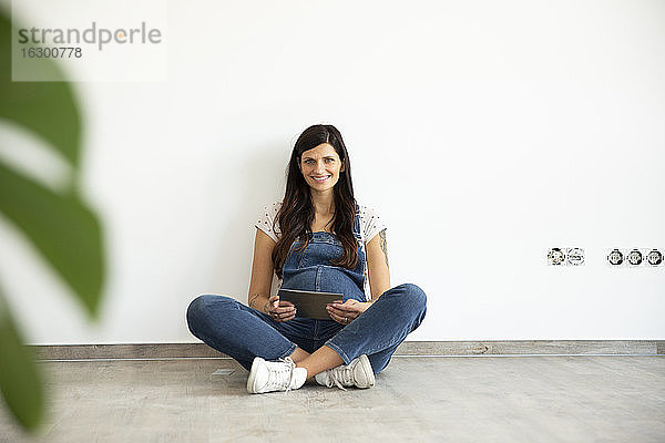 Lächelnde schwangere Frau  die ein digitales Tablet benutzt  während sie an einer weißen Wand in ihrem neuen Haus sitzt