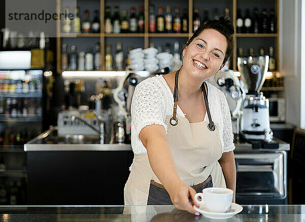 Lächelnde Besitzerin  die eine Kaffeetasse auf dem Tresen serviert  während sie in einem Café arbeitet