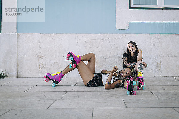Lächelnde Freundinnen mit Rollschuhen  die sich auf dem Fußweg vor einer Mauer in der Stadt entspannen