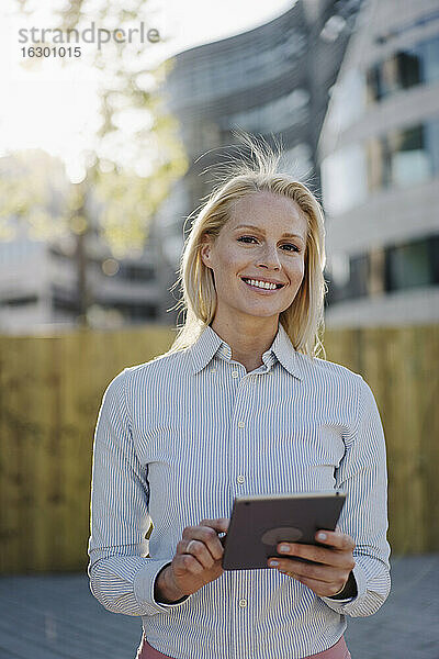 Lächelnde blonde Geschäftsfrau mit digitalem Tablet im Finanzviertel der Stadt