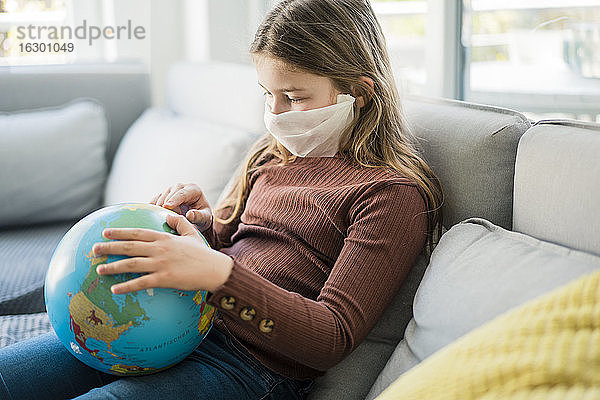 Mädchen liest Globus und trägt Gesichtsmaske auf Sofa zu Hause