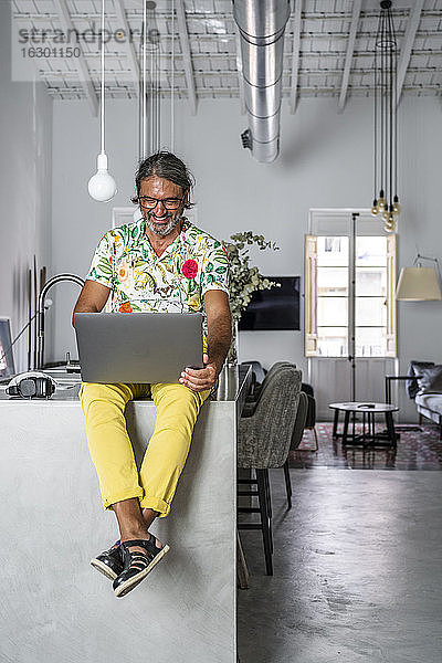Lächelnder Mann  der einen Laptop benutzt  während er zu Hause auf dem Tresen sitzt