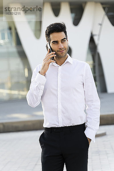 Selbstbewusster Geschäftsmann mit Hand in der Tasche  der über sein Smartphone spricht  während er in der Stadt steht