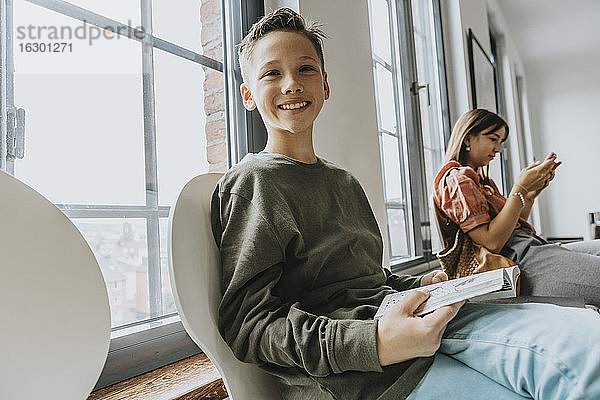 Lächelnder Junge mit Buch auf einem Stuhl sitzend  während ein Teenager-Mädchen im Wartezimmer ein Smartphone benutzt