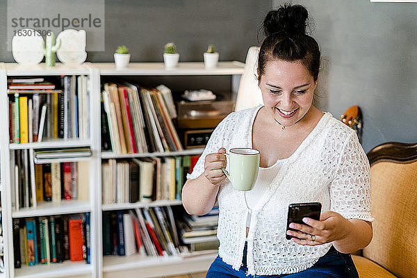 Lächelnde  üppige Frau  die einen Kaffeebecher hält und ein Smartphone benutzt  während sie in einem Café sitzt