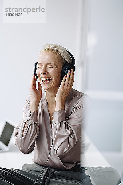 Fröhliche Geschäftsfrau hört Musik über Kopfhörer  während sie im Büro an der Wand sitzt