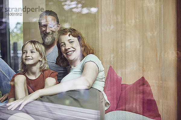 Lächelnde Eltern mit Tochter  die sich zu Hause auf dem Bett ausruhen  durch ein Fenster gesehen