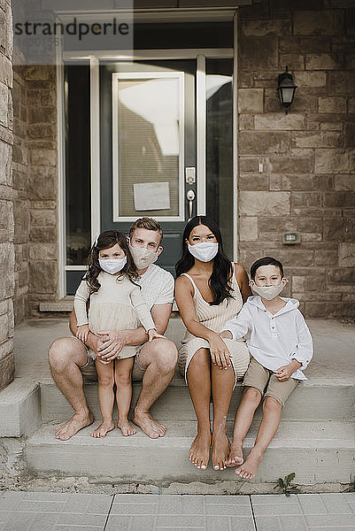 Familie mit Kindern trägt Schutzmaske  während sie auf den Stufen vor dem Haus sitzen
