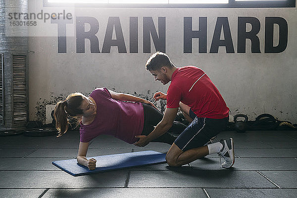 Fitnesslehrer  der einer Sportlerin beim Üben der Plank-Position auf einer Matte im Fitnessstudio hilft