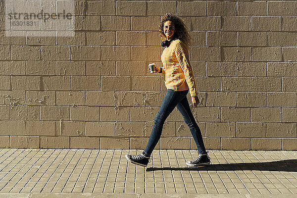Junge Frau  die einen Einwegbecher hält  während sie an einem sonnigen Tag auf einem Fußweg an einer Mauer entlang läuft