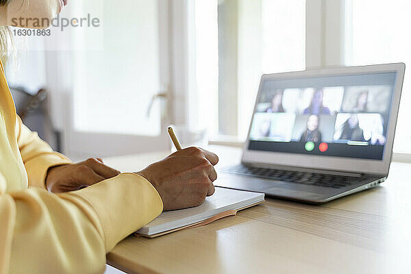 Abgeschnittene Hände einer Unternehmerin  die in ein Tagebuch schreibt  während sie mit Kollegen per Videokonferenz zu Hause spricht