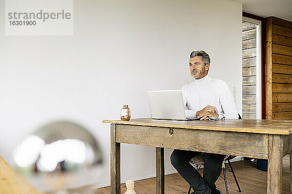 Nachdenklicher Geschäftsmann mit Laptop auf dem Schreibtisch sitzend an der Wand im Büro