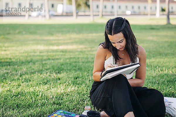 Frau malt in einem Buch  während sie im Gras eines öffentlichen Parks sitzt