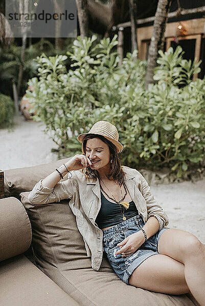 Lächelnde Frau mit Hut sitzt auf einer Couch im Freien