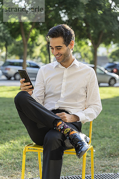 Lächelnder Geschäftsmann  der ein Mobiltelefon benutzt  während er auf einem Stuhl vor einem Baum sitzt