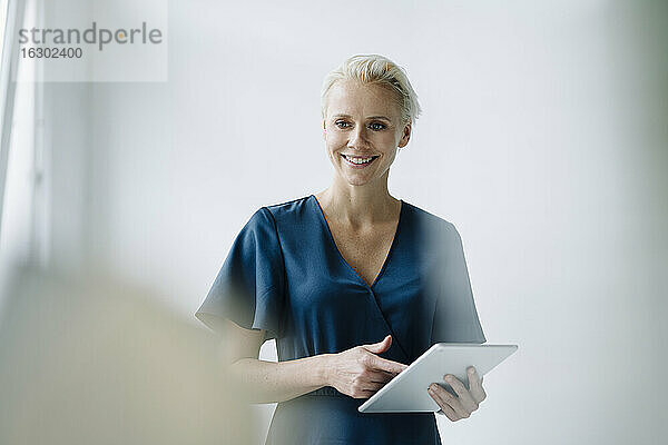 Weibliche Fachkraft  die ein digitales Tablet benutzt und wegschaut  während sie im Büro an der Wand steht