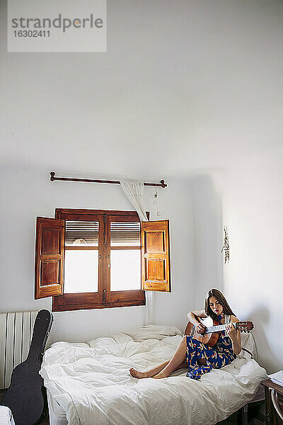 Junge Frau übt Gitarre  während sie zu Hause auf dem Bett sitzt