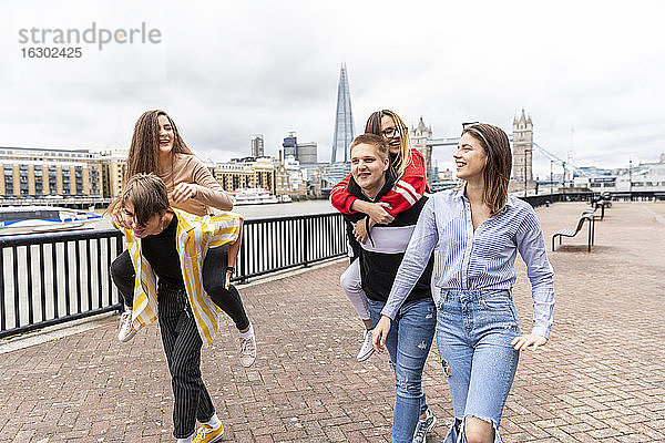 Männliche Freunde  die ihre Freundinnen beim Gehen auf einem Fußweg in der Stadt London huckepack nehmen  Großbritannien