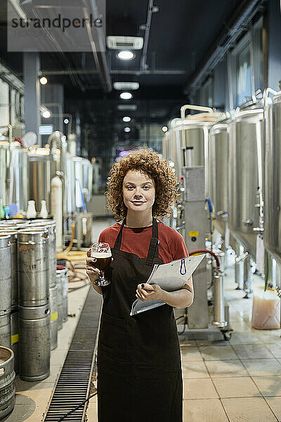 Porträt einer selbstbewussten Frau  die ein Klemmbrett und ein Bierglas in einer Handwerksbrauerei hält