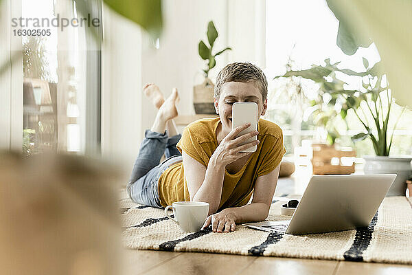 Frau  die ein Mobiltelefon benutzt  während sie mit einer Kaffeetasse und einem Laptop auf dem Teppich zu Hause liegt