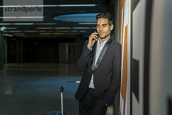 Männlicher Berufstätiger mit Koffer  der über sein Smartphone spricht  während er an der Wand eines Bahnhofs steht