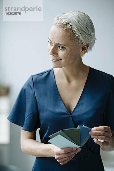 Lächelnde Geschäftsfrau  die ein Farbmuster hält und wegschaut  während sie im Büro steht