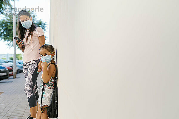 Frau mit Gesichtsmaske  die ein Mobiltelefon benutzt und ein Mädchen betrachtet  das sich an eine weiße Wand lehnt