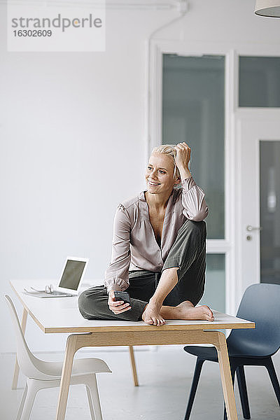 Nachdenkliche Unternehmerin  die ihr Smartphone in der Hand hält  während sie auf dem Schreibtisch an der Wand im Büro sitzt