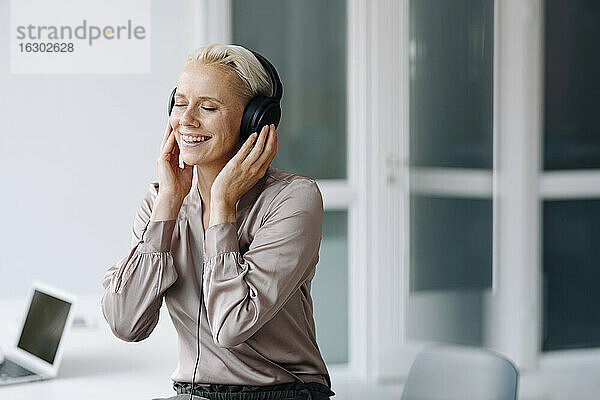 Lächelnde Geschäftsfrau hört Musik über Kopfhörer  während sie im Büro sitzt