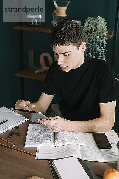 Gut aussehender Mann  der ein digitales Tablet benutzt  während er am Tisch sitzt und Hausaufgaben macht